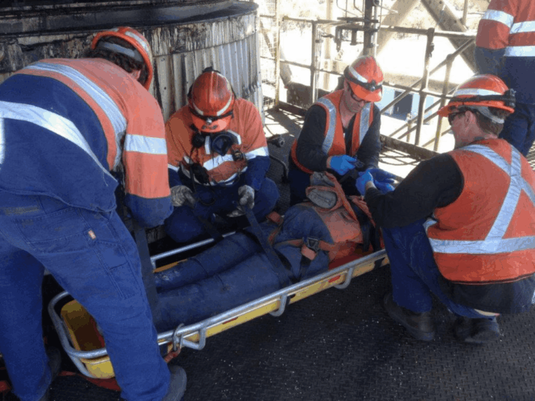 Trained workers providing emergency first aid and safely securing an injured person on a stretcher at an industrial worksite.